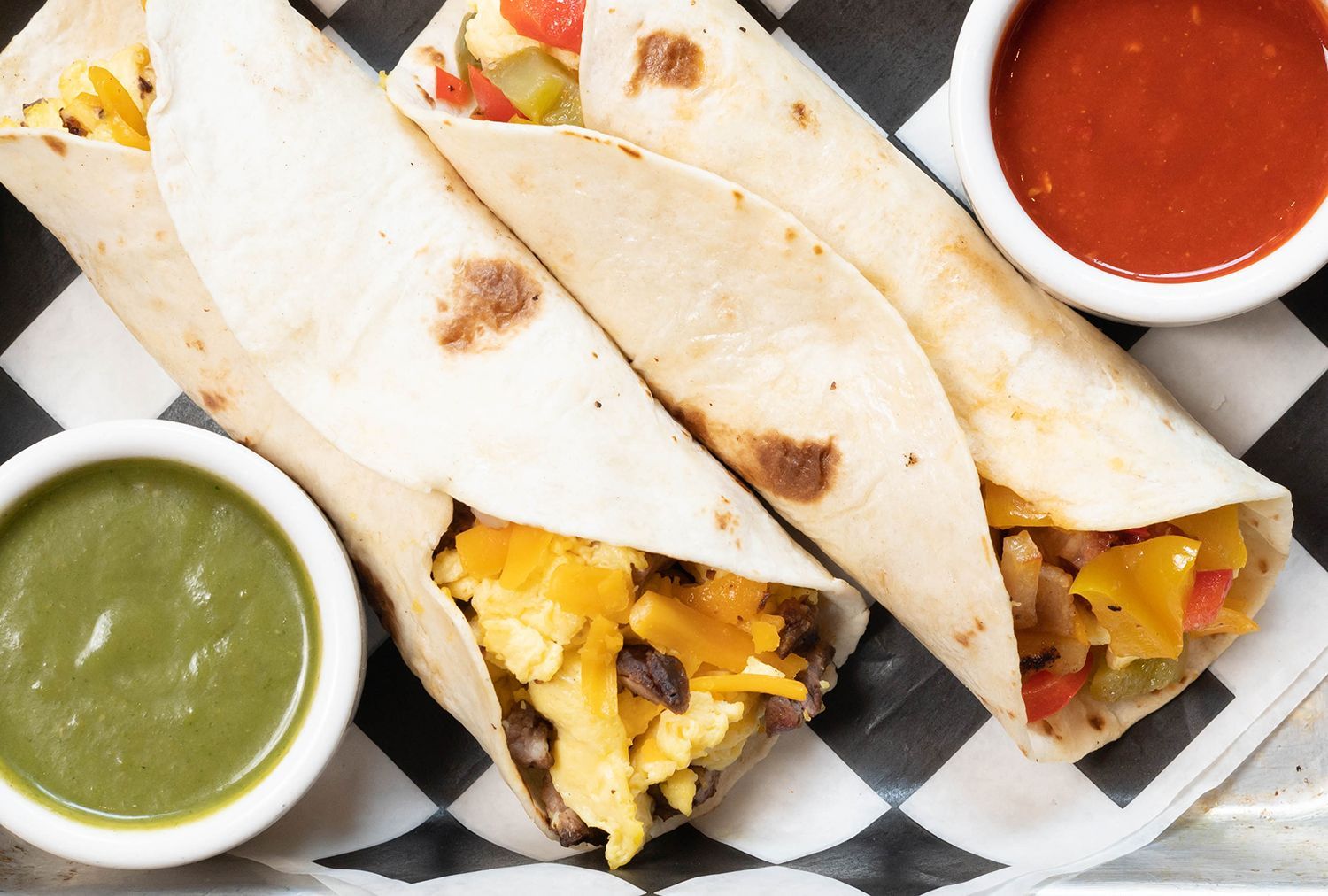 Three burritos are sitting on a checkered paper next to two bowls of sauce.