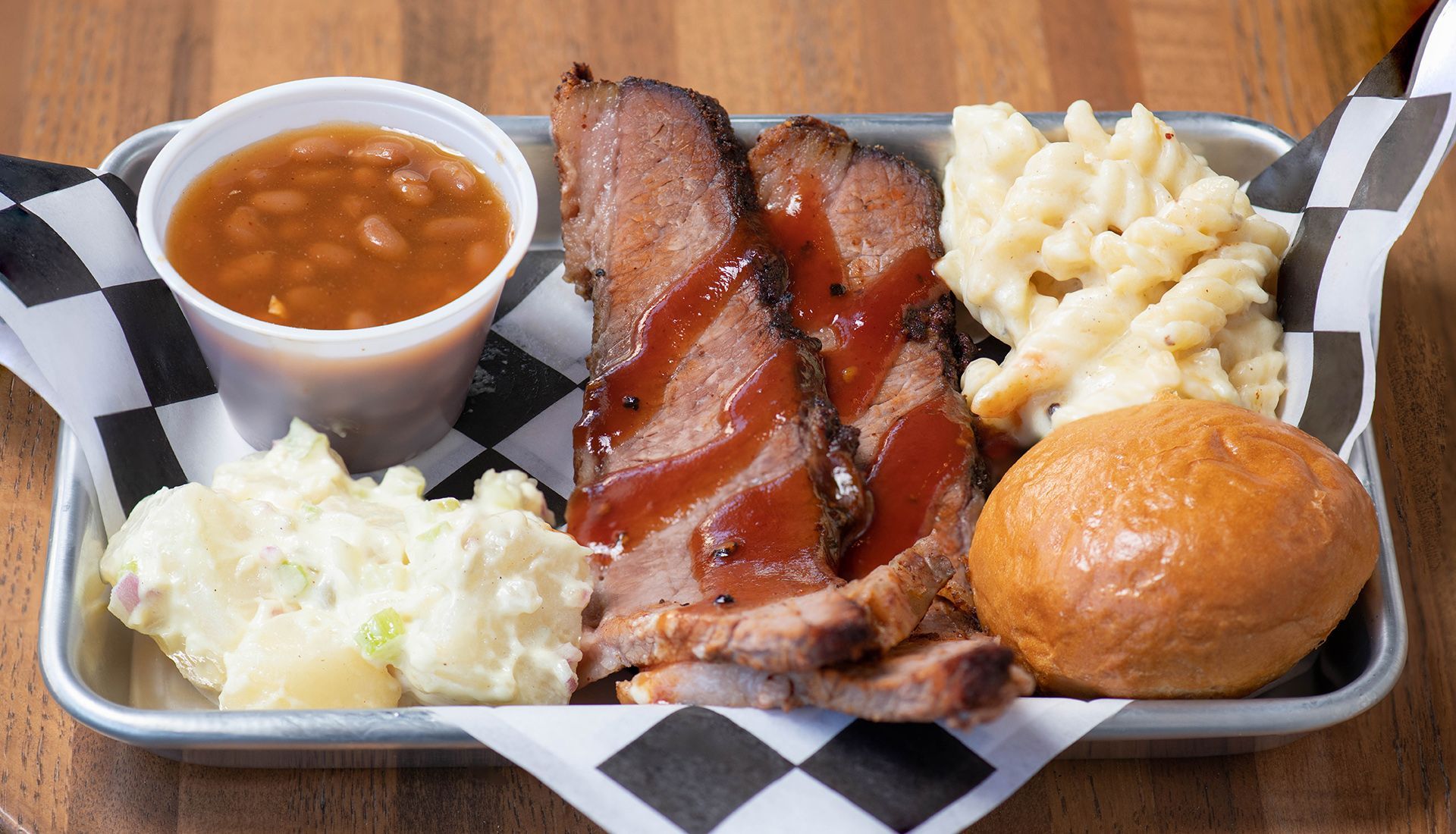 A tray of food with meat , beans , mashed potatoes and a roll on a wooden table.