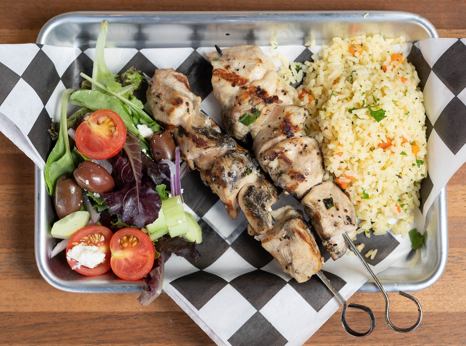 A tray of food with chicken skewers , rice and salad on a checkered napkin.