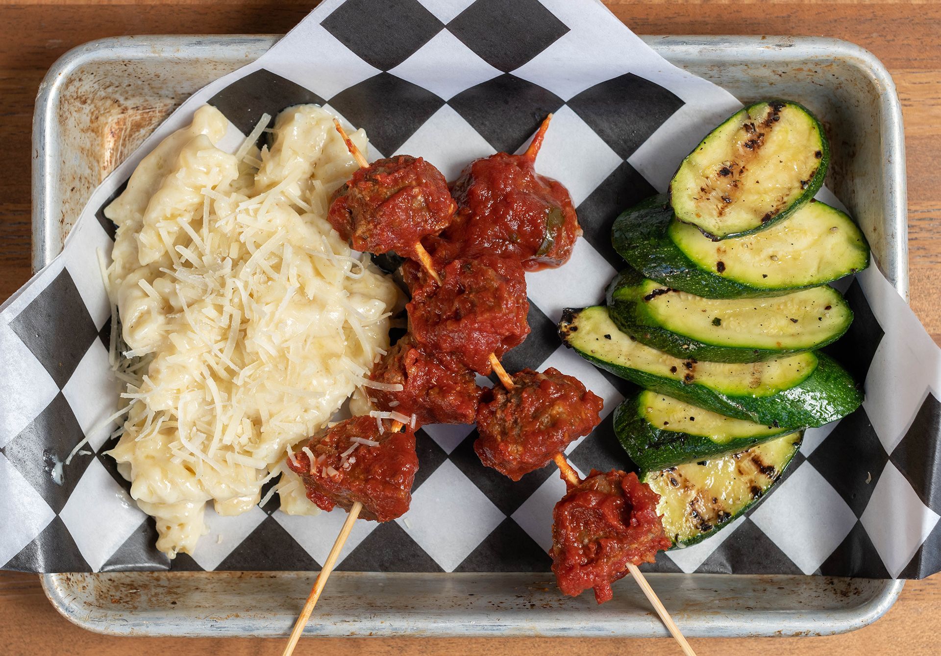 A tray of food with meatballs and vegetables on skewers on a checkered paper.