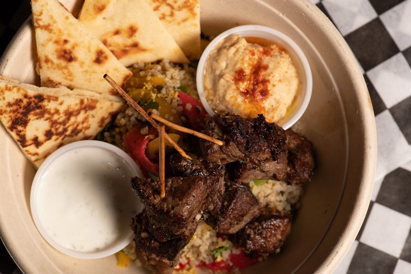 A bowl of food with meat , rice , pita bread and dips on a checkered table cloth.