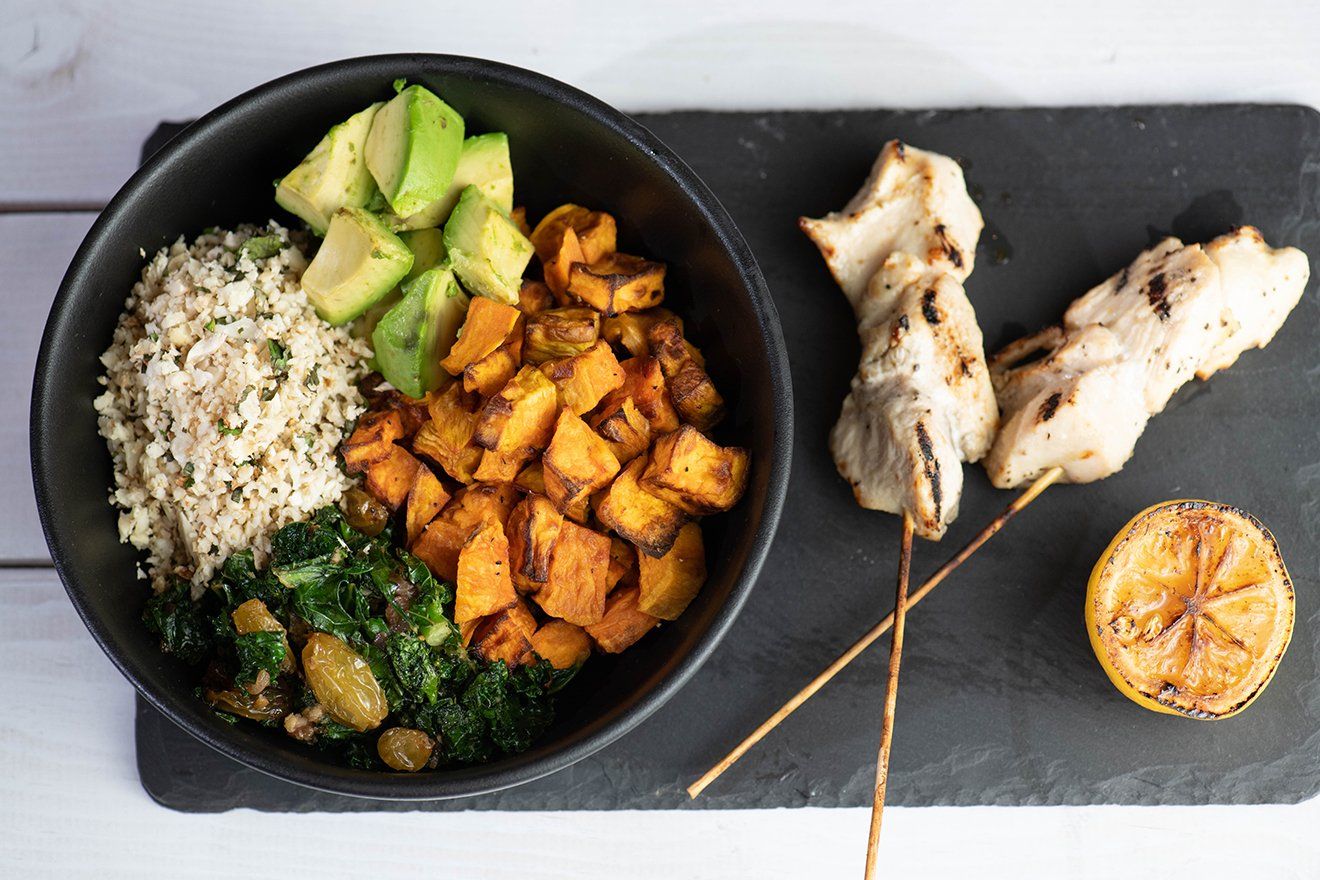 A bowl of food with rice , sweet potatoes , avocado and chicken skewers on a cutting board.