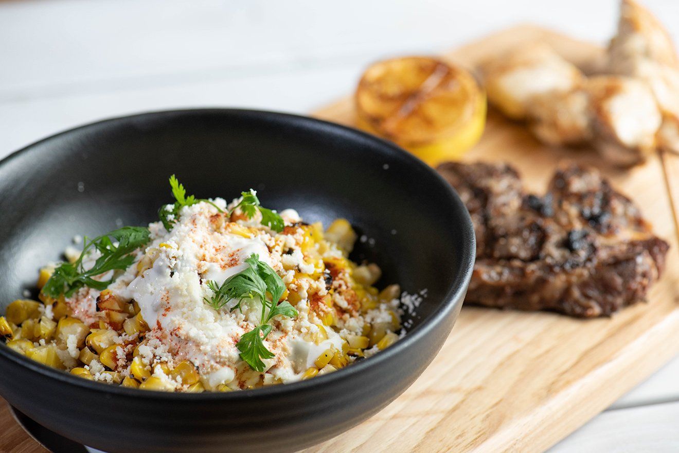 A bowl of corn on the cob is sitting on a wooden cutting board next to a steak.