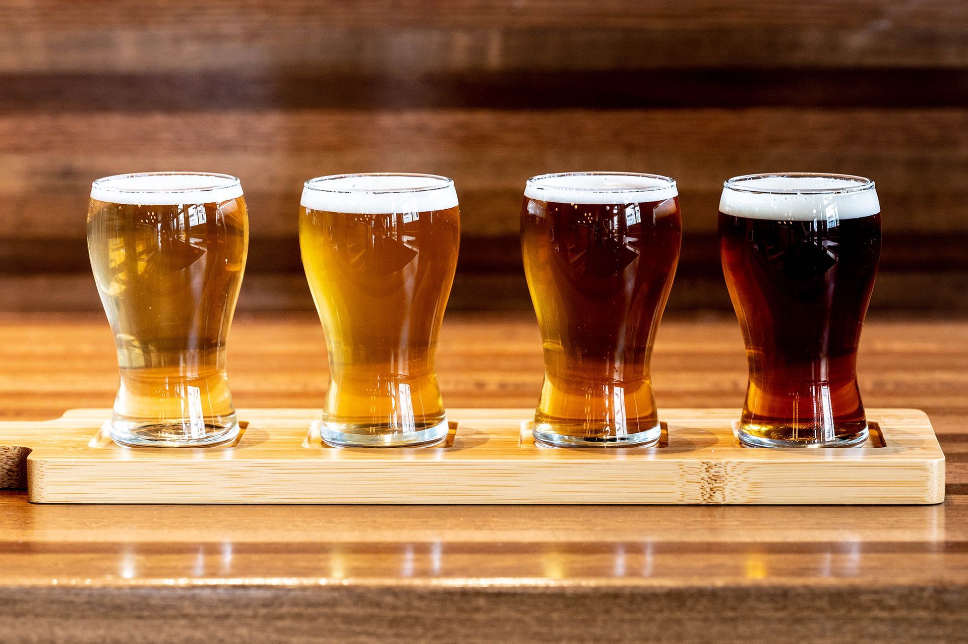 Four glasses of beer are lined up on a wooden tray on a wooden table.