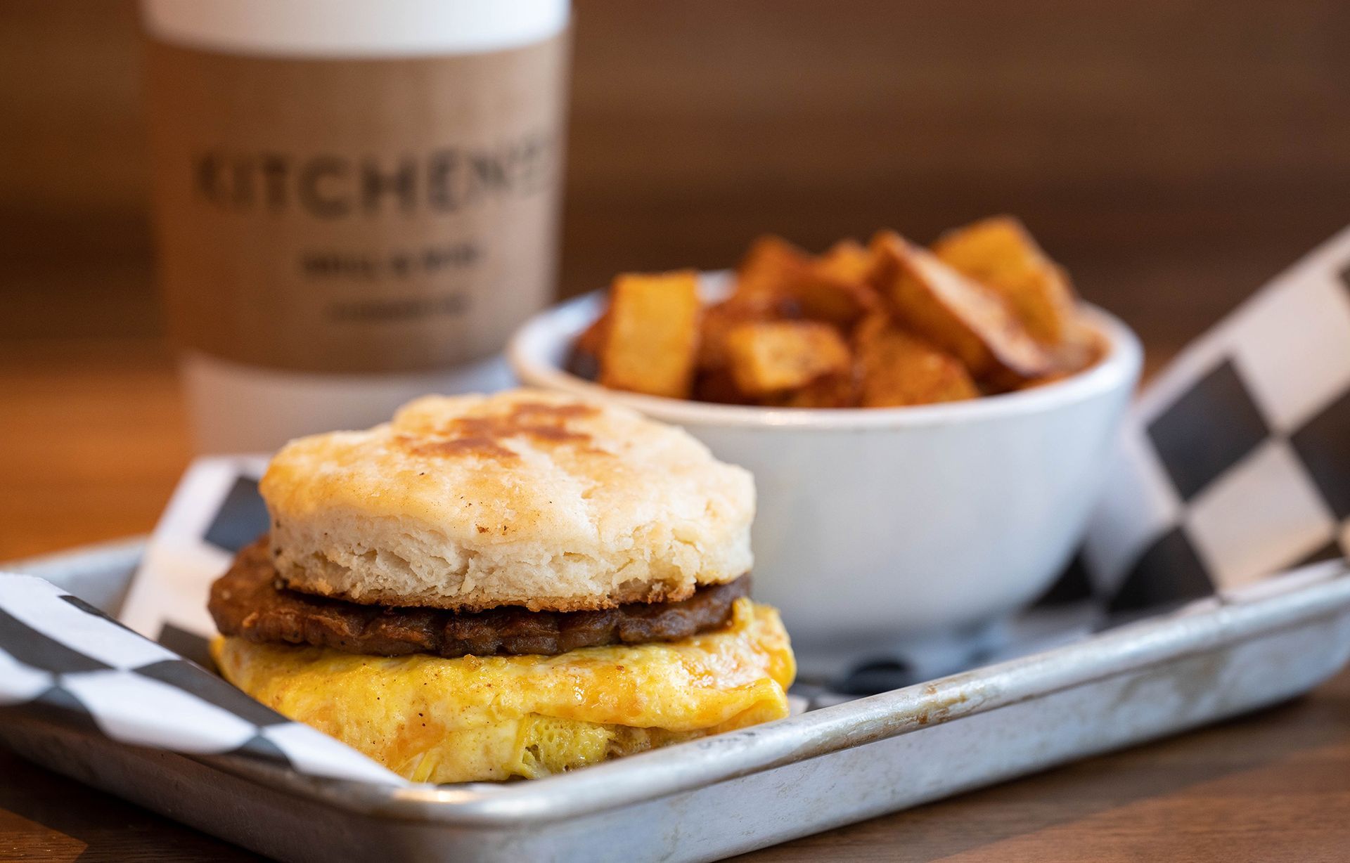 A breakfast sandwich with eggs and sausage on a biscuit on a tray next to a bowl of potatoes.