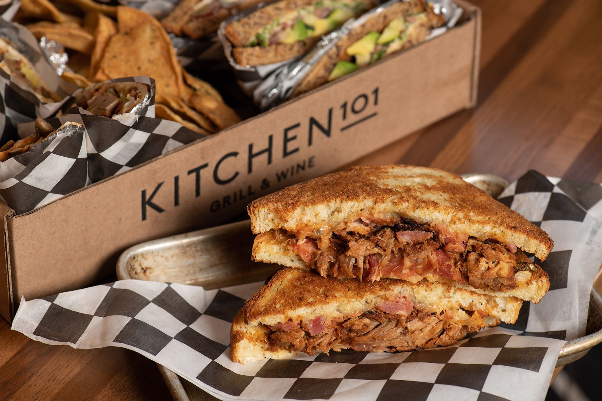 A sandwich is sitting on a checkered napkin next to a box of food.