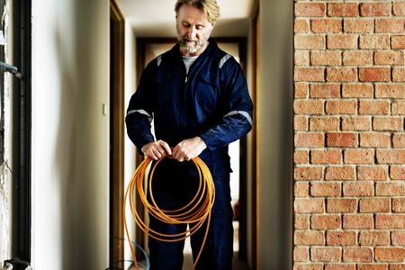 A man in a blue jumpsuit is holding a coil of orange wire.