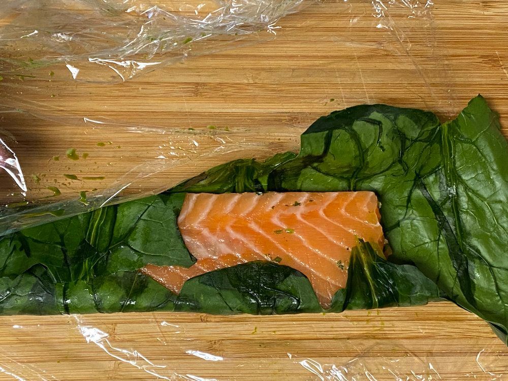 A piece of salmon wrapped in spinach leaves on a wooden cutting board.