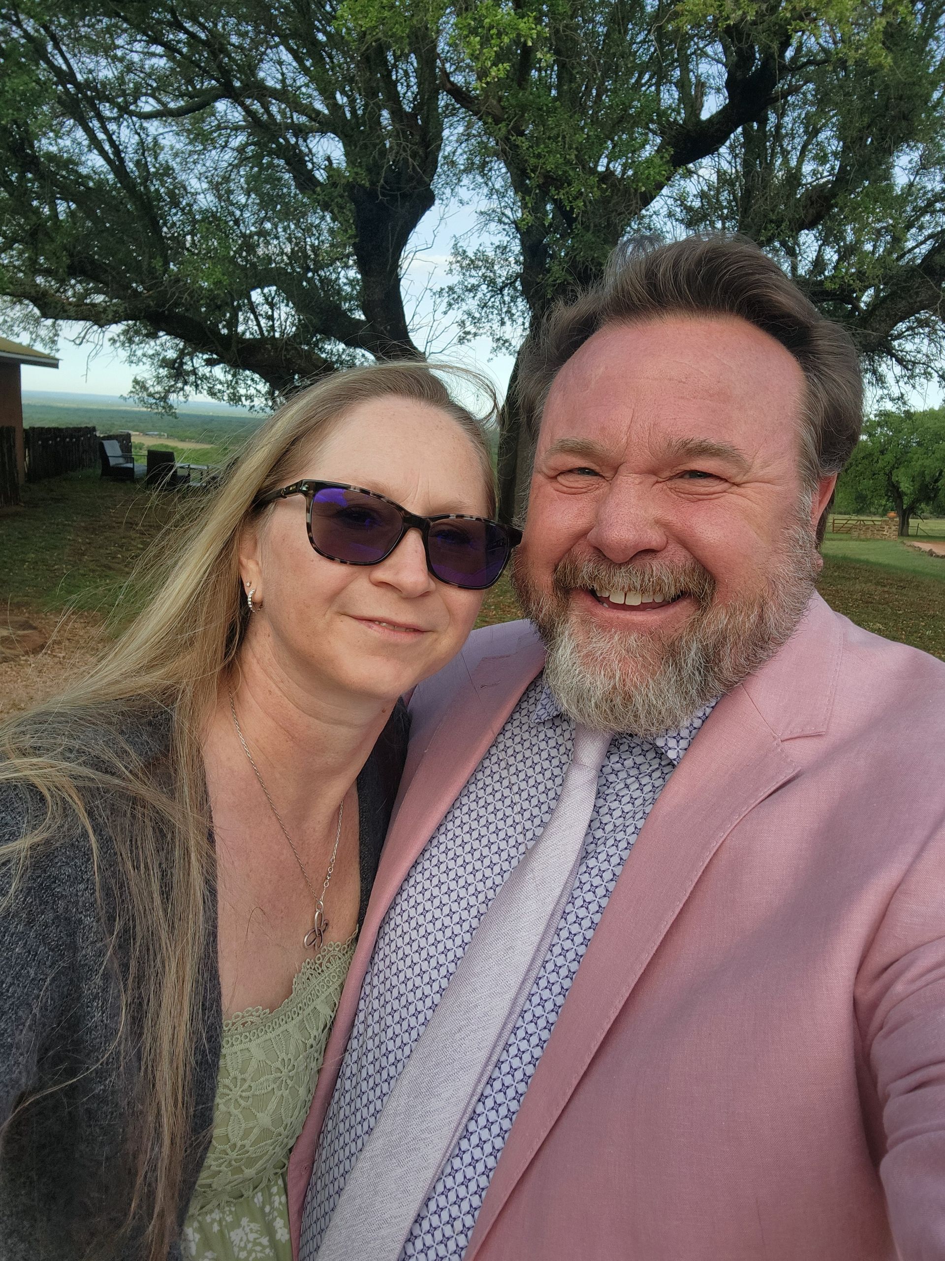 A smiling couple in an outdoor setting, man in pink jacket, woman in sunglasses, both close together.