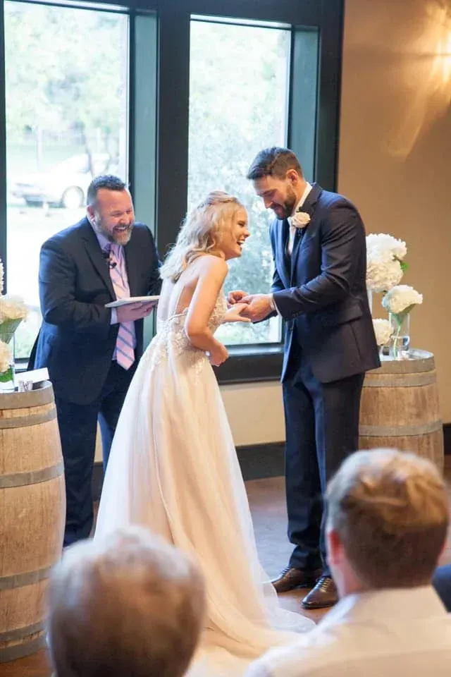 Groom placing ring on bride's finger during a wedding ceremony indoors, officiant laughing, barrels used as decor.