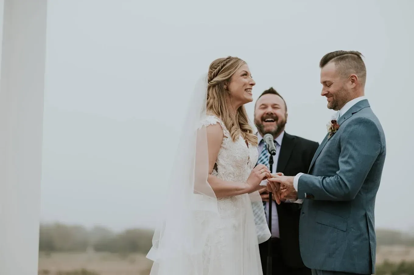 Couple exchanging rings during an outdoor wedding ceremony; officiant smiles.