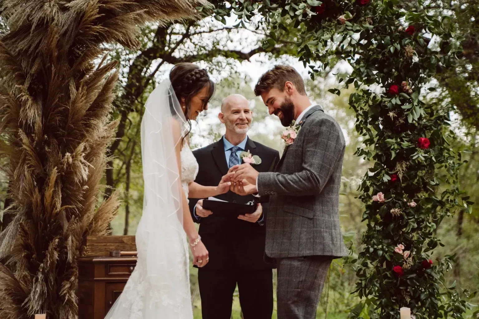 Wedding ceremony: Couple exchanging rings under archway decorated with pampas grass and flowers, officiant smiling.