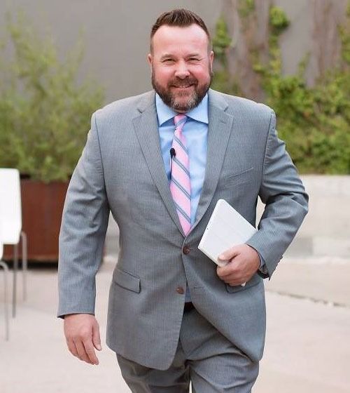 James Simmons in grey suit smiles and walks, holding a white tablet outdoors. Blue tie with pink stripes.
