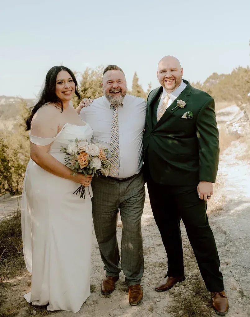 Bride and two men posing outdoors; bride in white dress, men in suit and button-down.