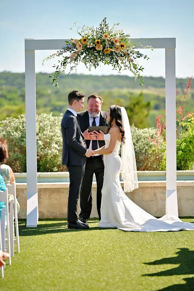 Couple exchanging vows under floral arch, officiant present. Outdoor ceremony on sunny day.