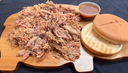 A pulled pork sandwich is on a wooden cutting board next to a cup of barbecue sauce.