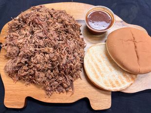 A wooden pig shaped cutting board with pulled pork , a sandwich and a cup of sauce.