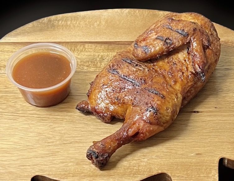 A grilled chicken is sitting on a wooden cutting board next to a small cup of sauce.