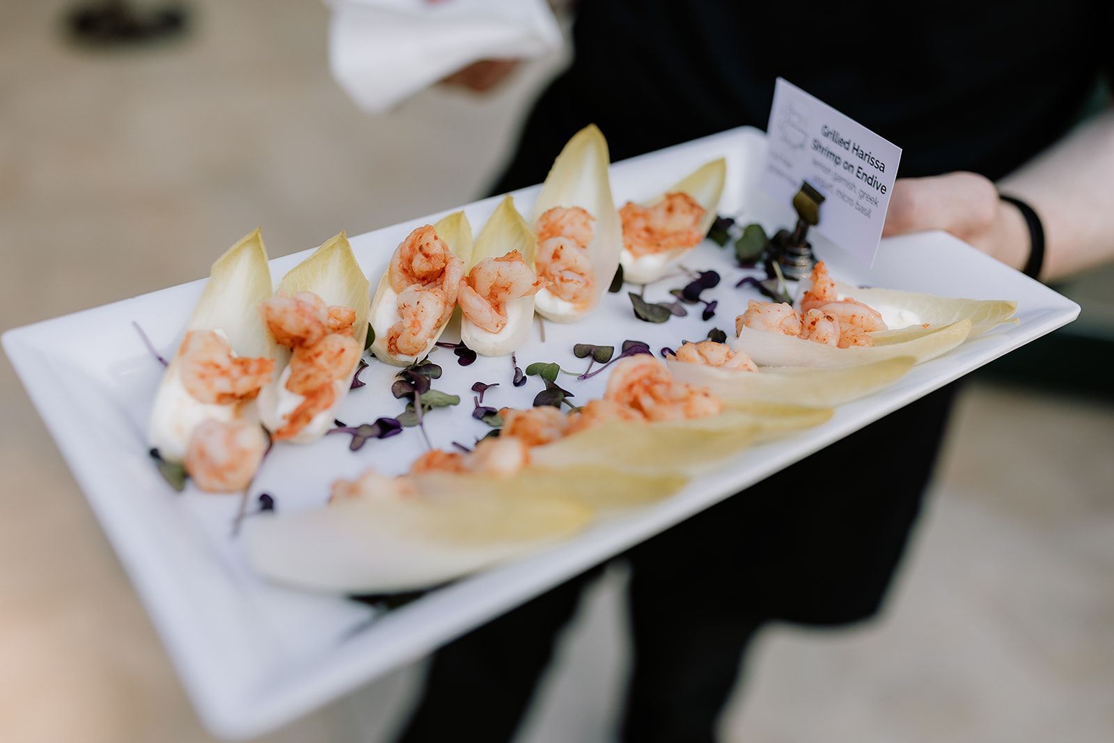 A server holds a white rectangular platter featuring several endive leaves topped with shrimp, cream, and microgreens.