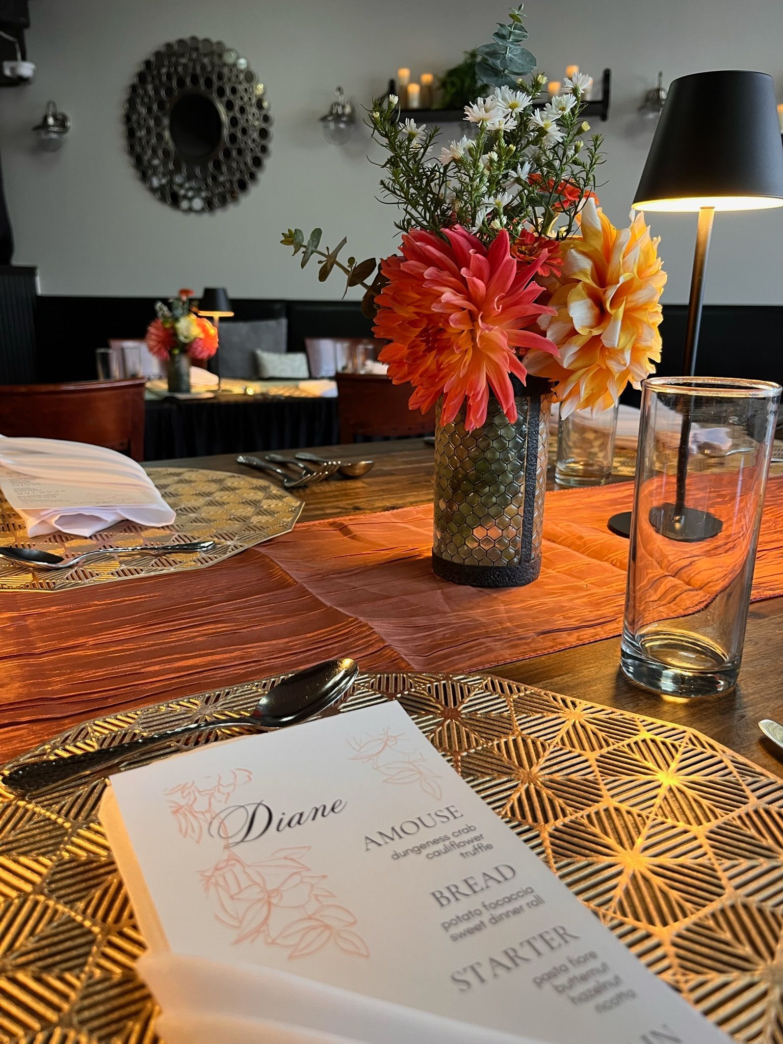 Table setting with floral centerpiece, menu, and glassware. Warm colors of orange and gold.