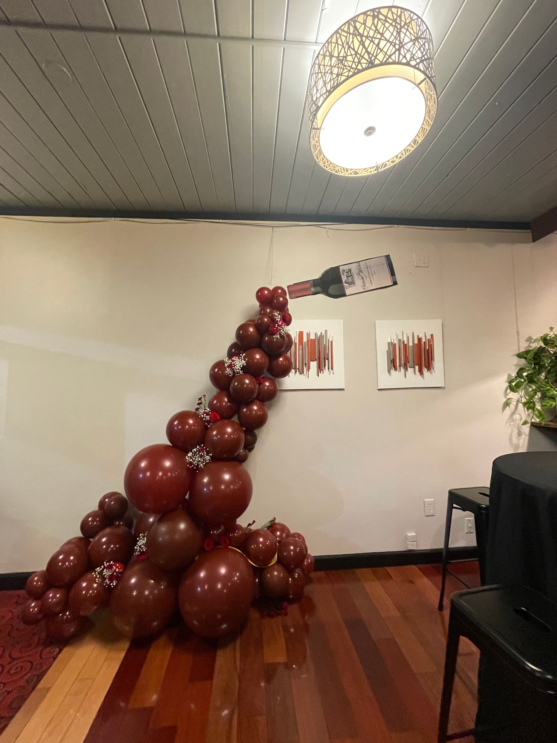 Balloon wine bottle cascade, red and brown balloons, on wooden floor. Art, chairs, and a light fixture.