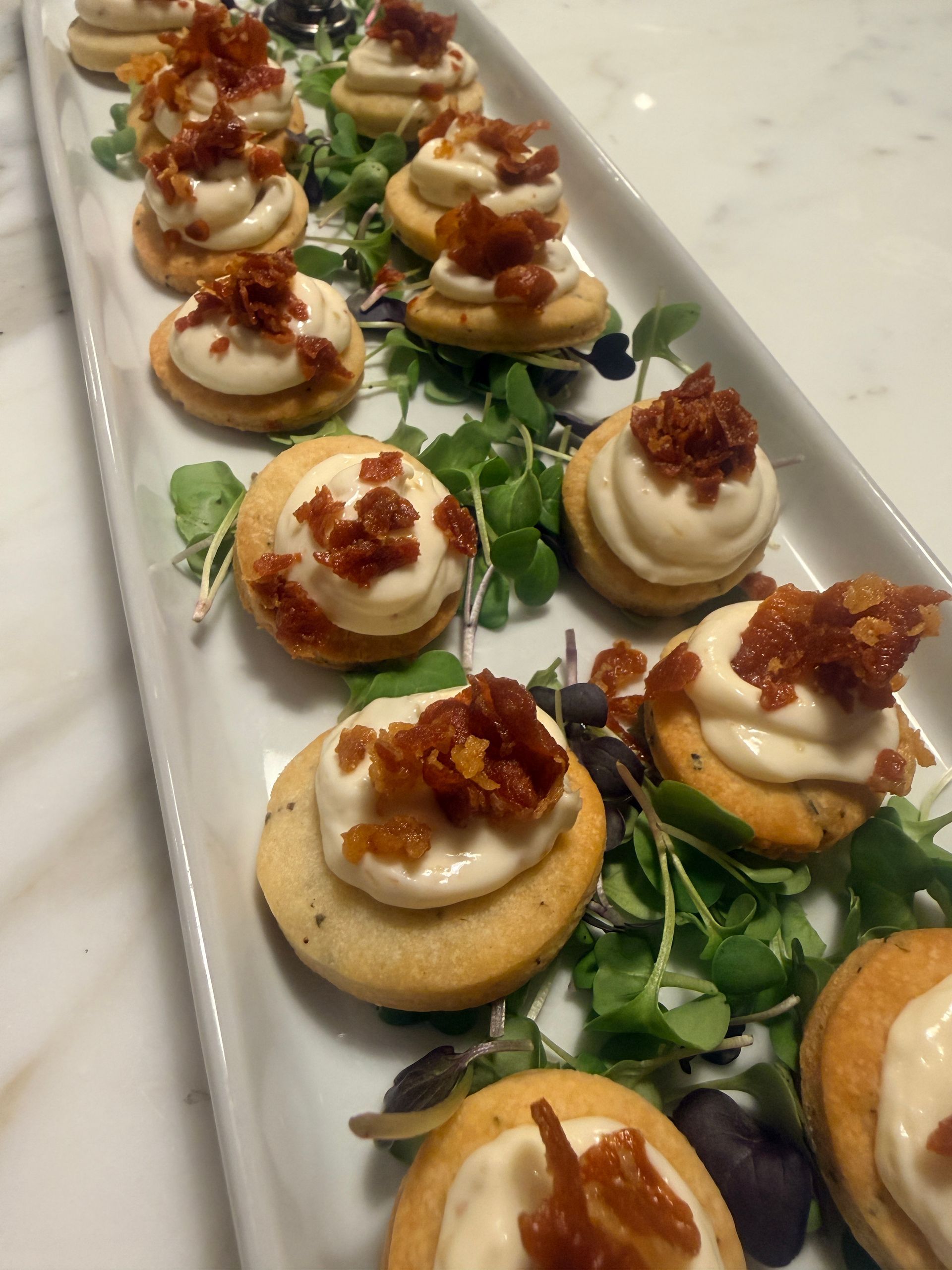 Appetizers on a white platter: round crackers topped with a white cream and crispy, brown bits, garnished with greens.