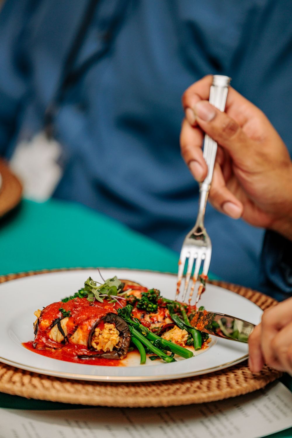 Person cutting into a plate of food with a fork and knife. Green beans and sauce visible.
