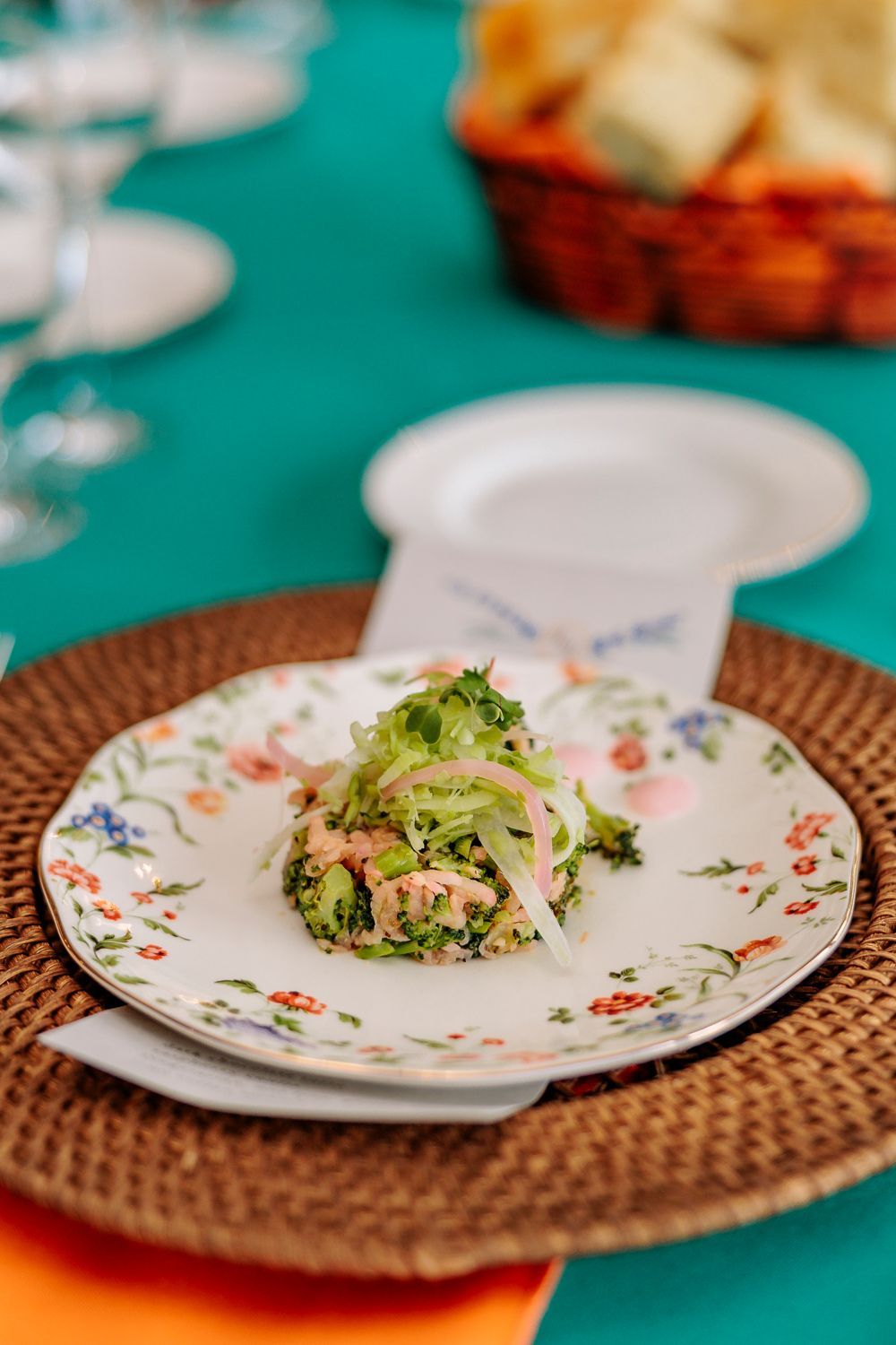 Salad on a floral plate atop a woven mat, set on a teal and orange table.