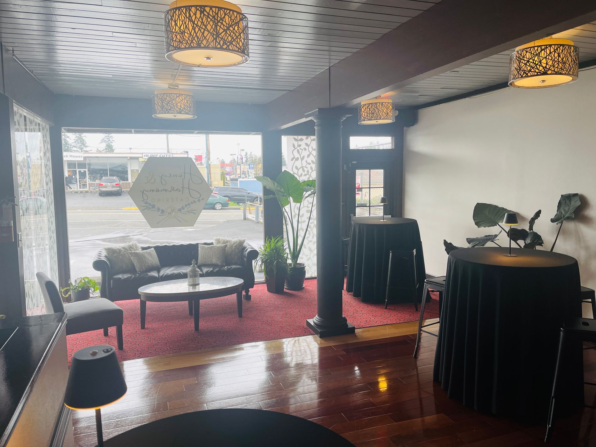Interior view of a venue with seating, round tables, large windows, decorative lighting, and plants.