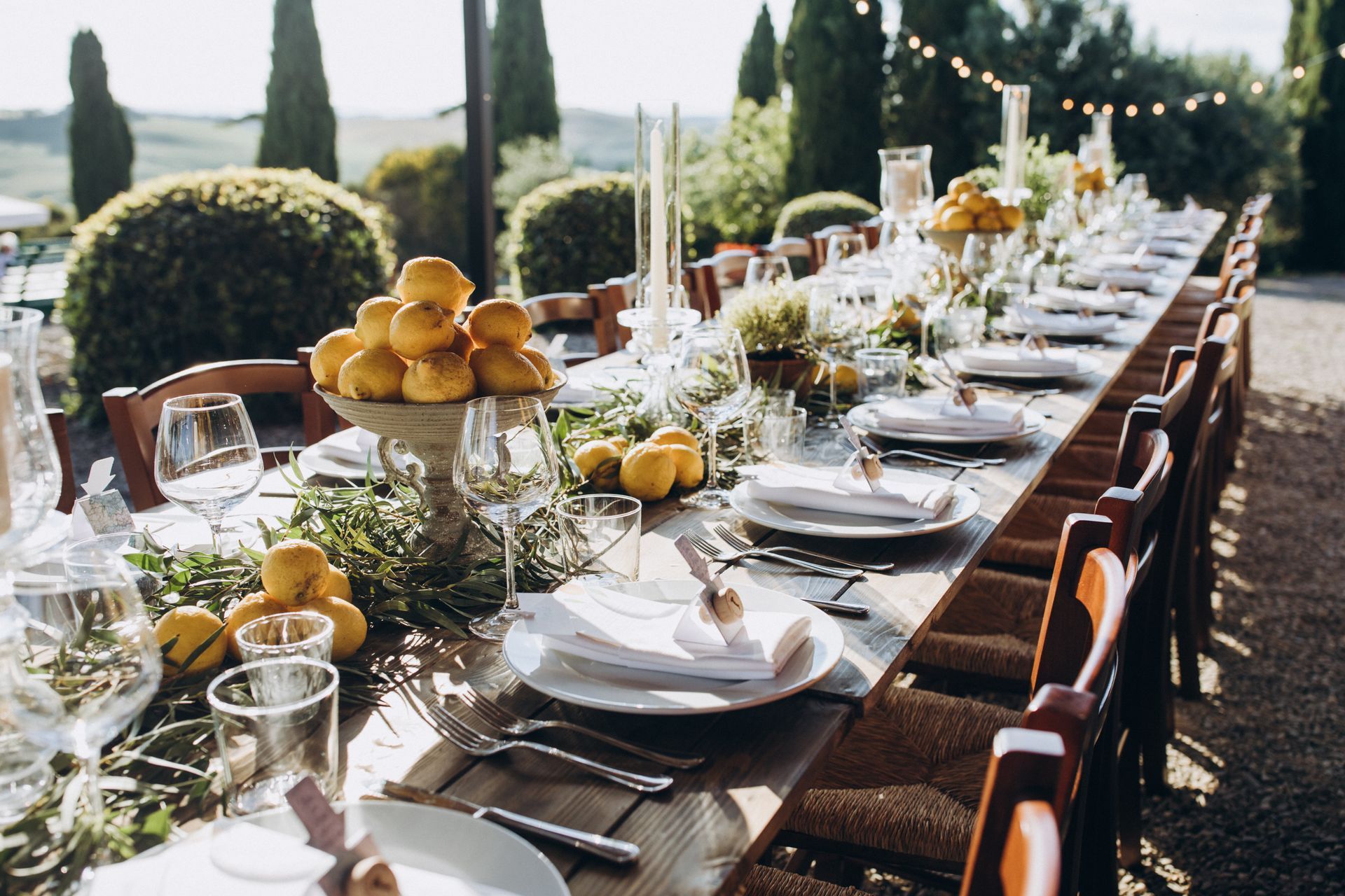 Long outdoor banquet table set with white dishes, flowers, and fruit in a garden