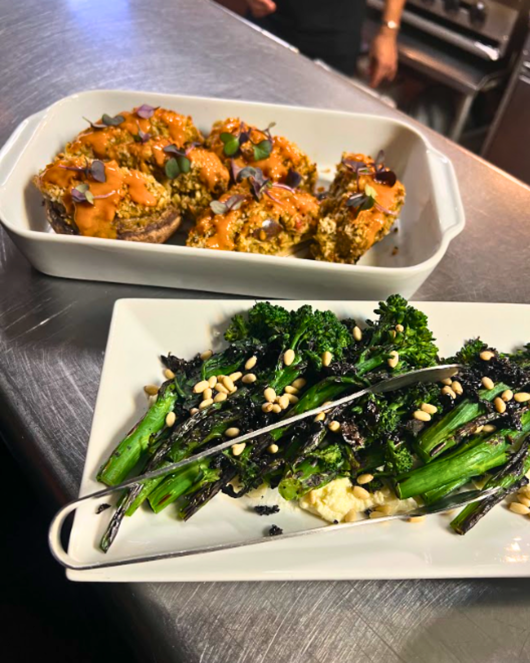 Two restaurant dishes: baked mushrooms and broccolini on a stainless steel surface.