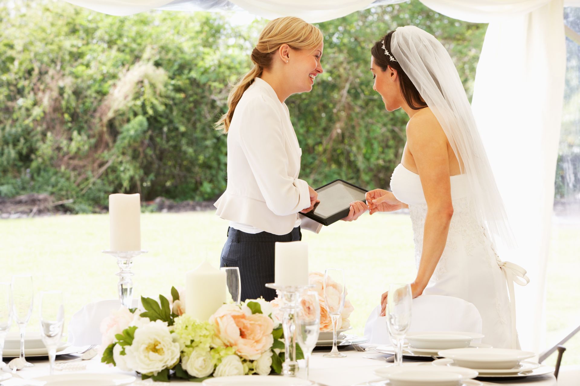 Bride in a wedding dress talks with a woman holding a tablet at a decorated table in a tent.