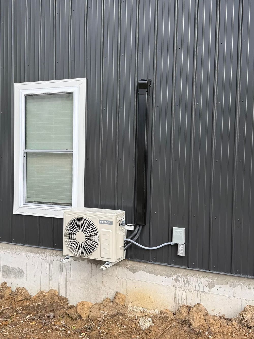 An outdoor mini-split heat pump unit mounted on a dark metal wall next to a window and an electrical disconnect box.