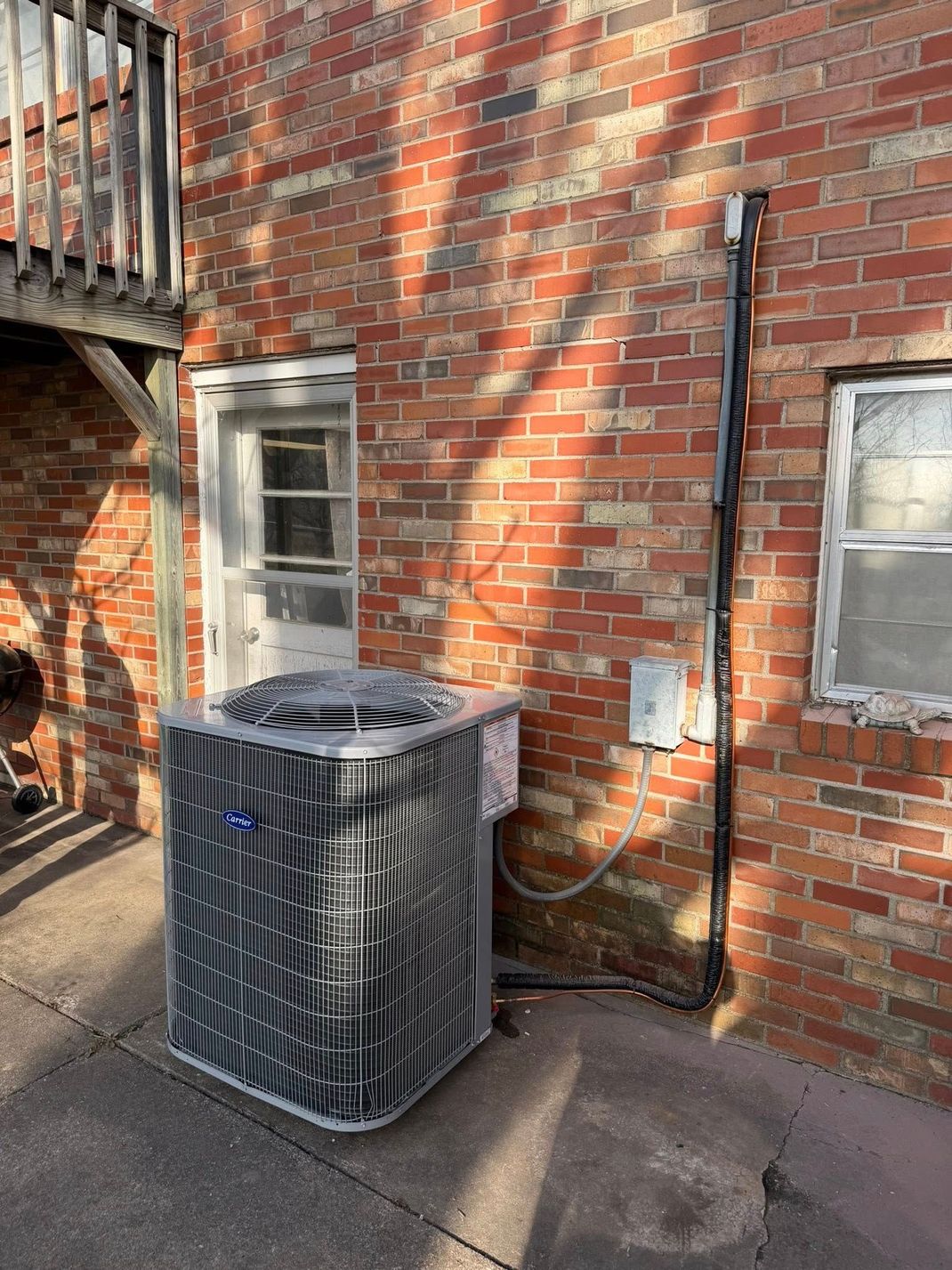 An air conditioning condenser sits outside on a concrete patio next to a brick building with a door and a window.