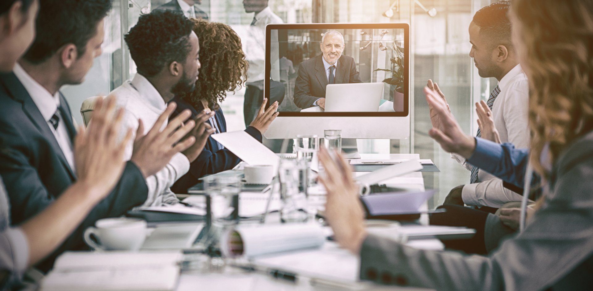 A group of business people are sitting around a table clapping their hands in front of a computer screen.