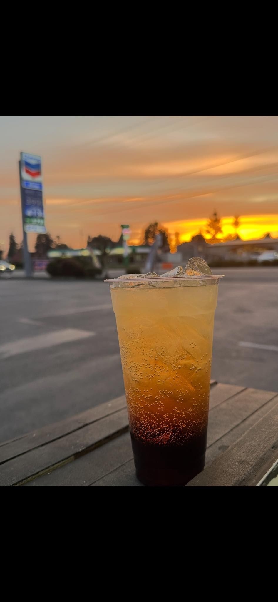A glass of soda is sitting on a wooden table in front of a sunset.