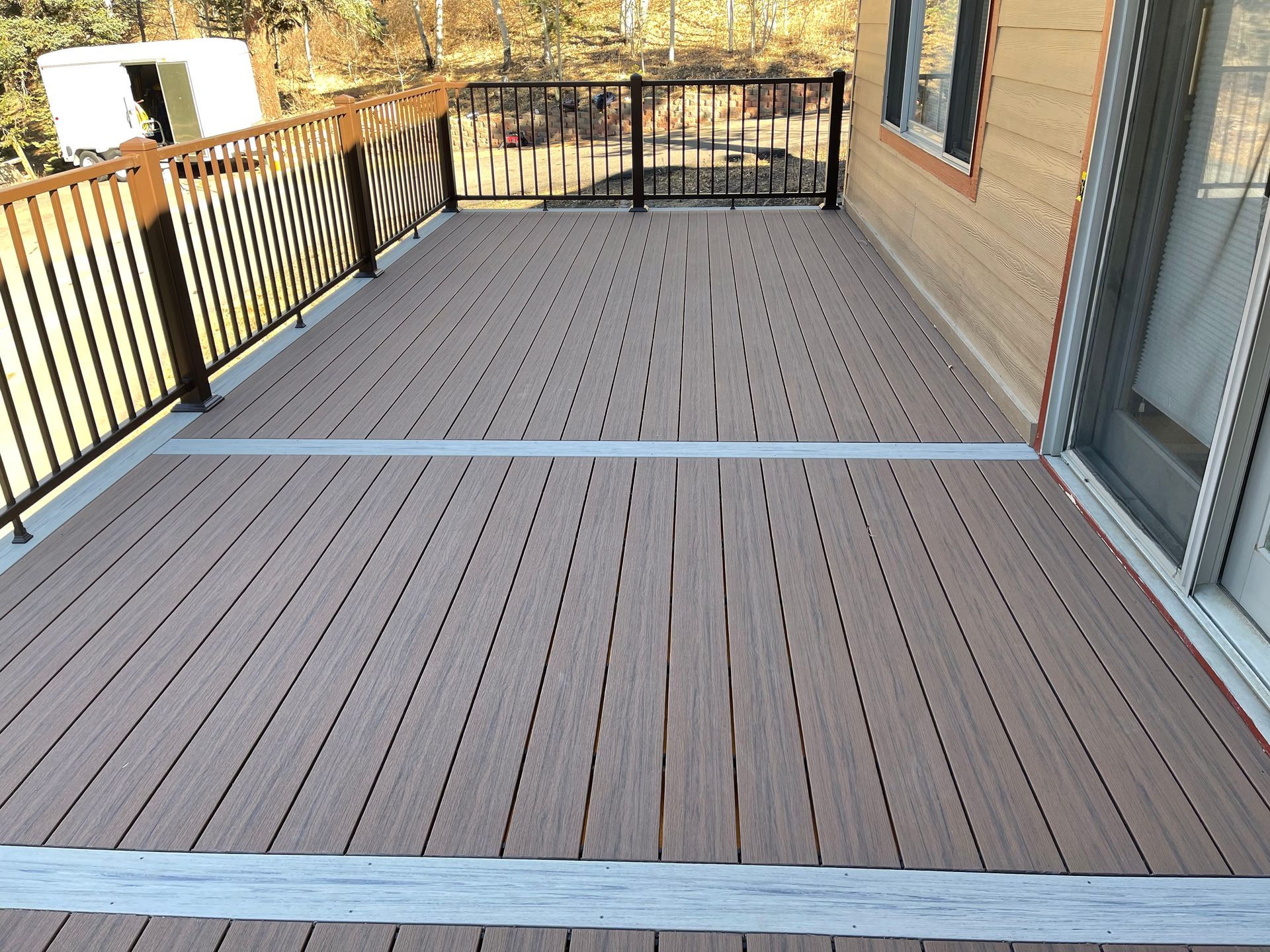 A spacious composite deck with light brown, wood-like planks and metal strips, bordered by a black metal railing, attached to a beige house with large windows and a sliding glass door. The deck is elevated, with a view of a backyard featuring trees and other natural elements in the background.