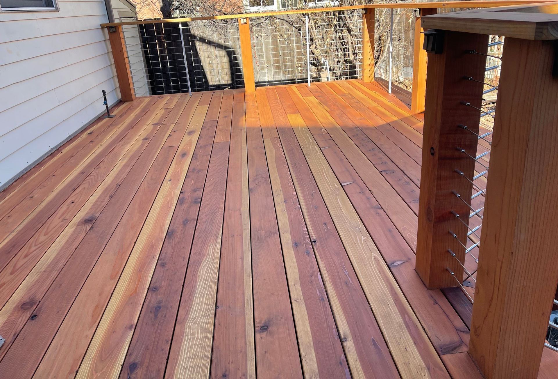 A wooden deck with rich, reddish-brown planks and a modern railing featuring horizontal metal cables and wooden posts. The deck is attached to a light-colored house, with a partial view of the backyard featuring trees and other natural elements in the background. Sunlight casts shadows across the deck, highlighting the natural grain and color variation of the wood.