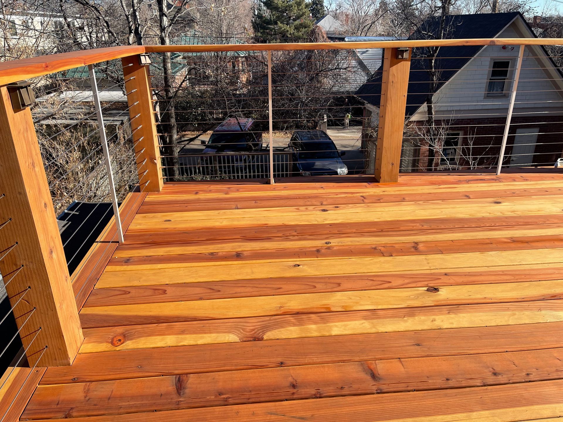 A beautifully refinished wooden deck with a modern cable railing system. The deck features rich, warm tones and a smooth, polished surface, overlooking a residential neighborhood with trees and houses in the background.