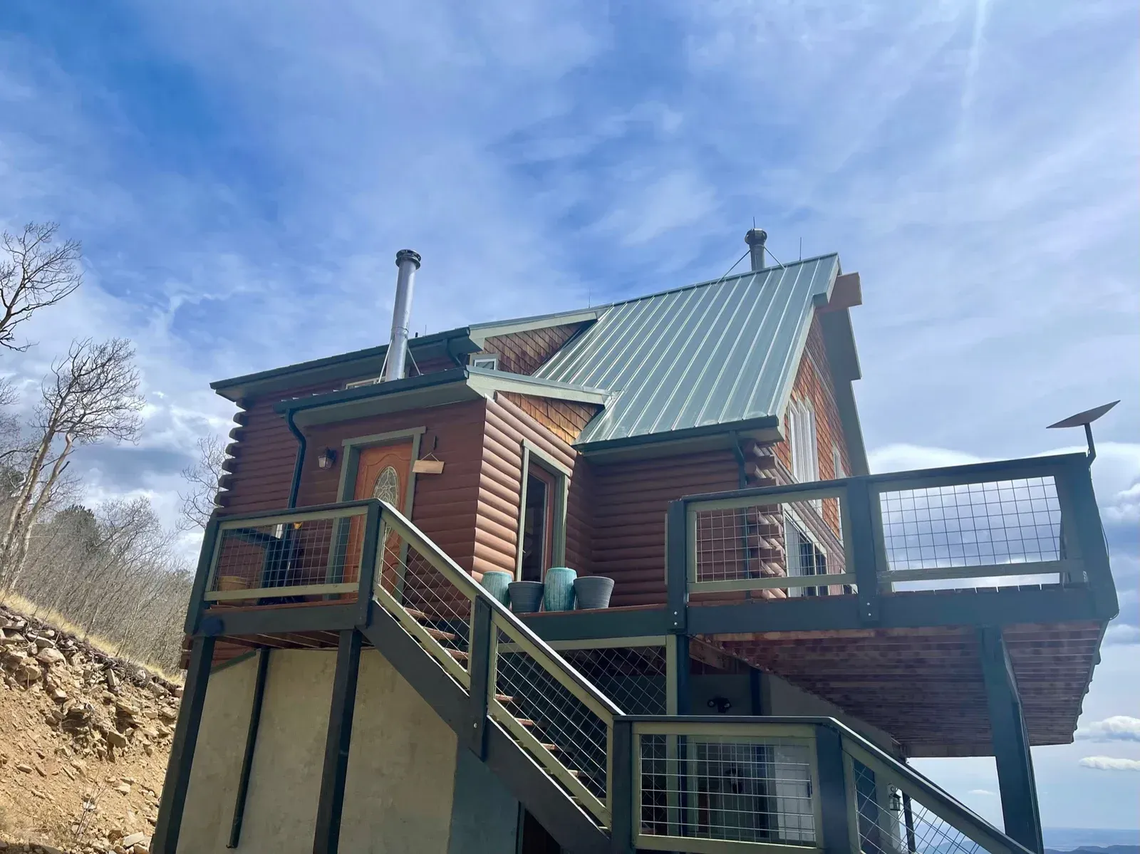 “Before photo of original cabin deck with green metal roof and simple wood railing on a mountain hillside home in Colorado.”