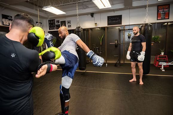 A man practices a kick on a pad held by another man in a gym.