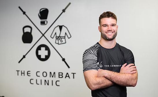 Man with arms crossed smiles in a gym. Sign for 
