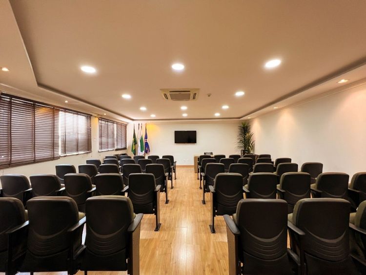 Sala de conferências vazia com fileiras de cadeiras pretas, piso de madeira, TV fixada na parede e bandeiras perto da entrada.