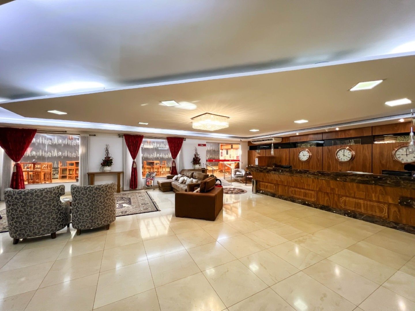Amplo salão de hotel com cadeiras estampadas, sofás, cortinas vermelhas e um bar de madeira.
