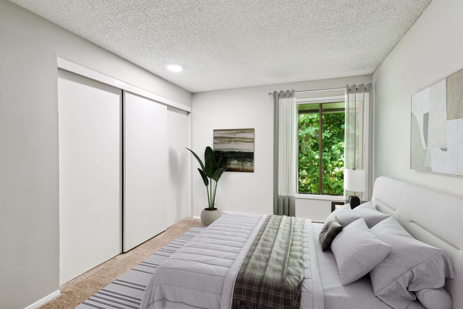 Bedroom in an apartment with white sliding closet doors, a bed, plant, and a window with sheer curtains.