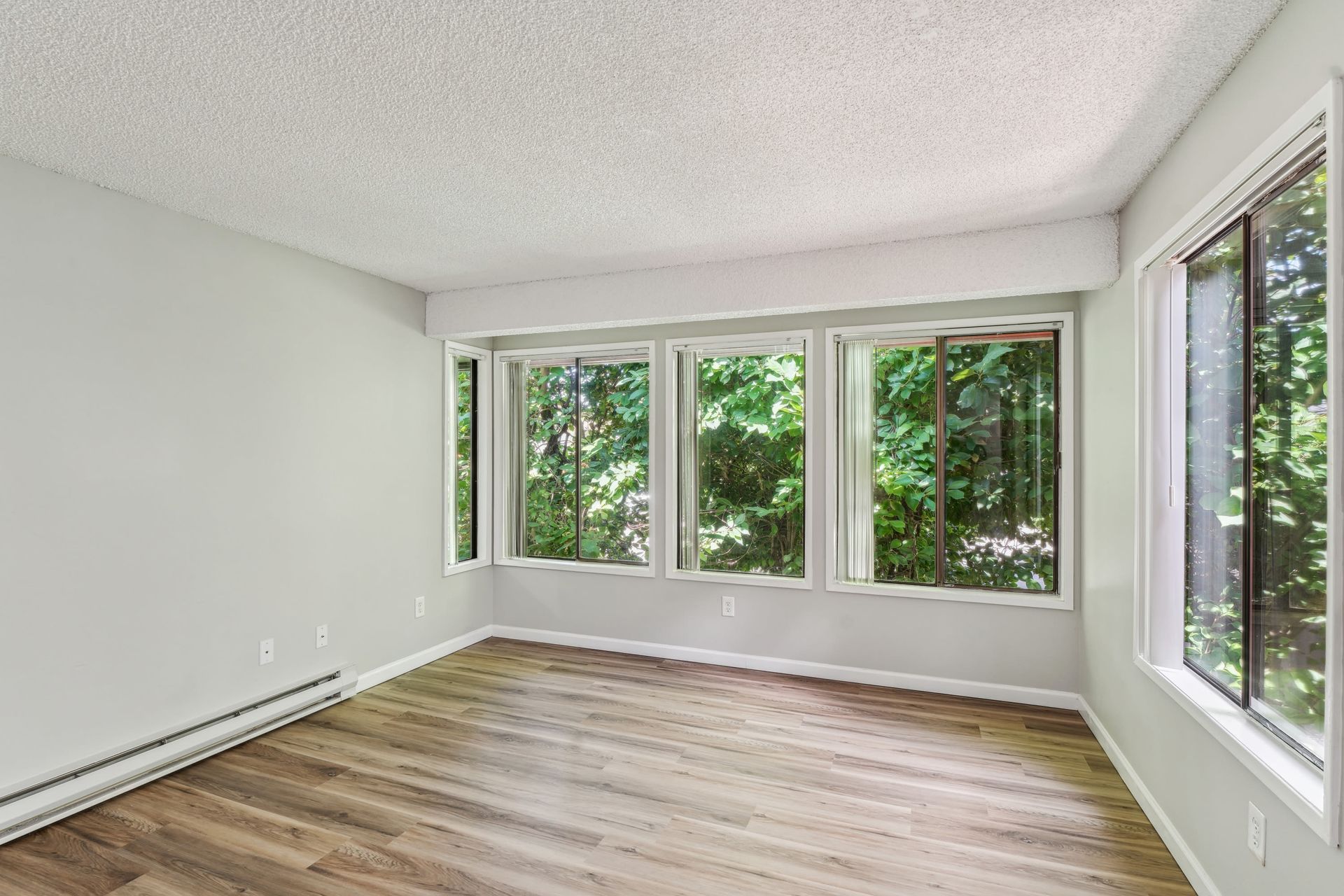 Bright empty living room with large windows facing greenery, wood-look flooring.
