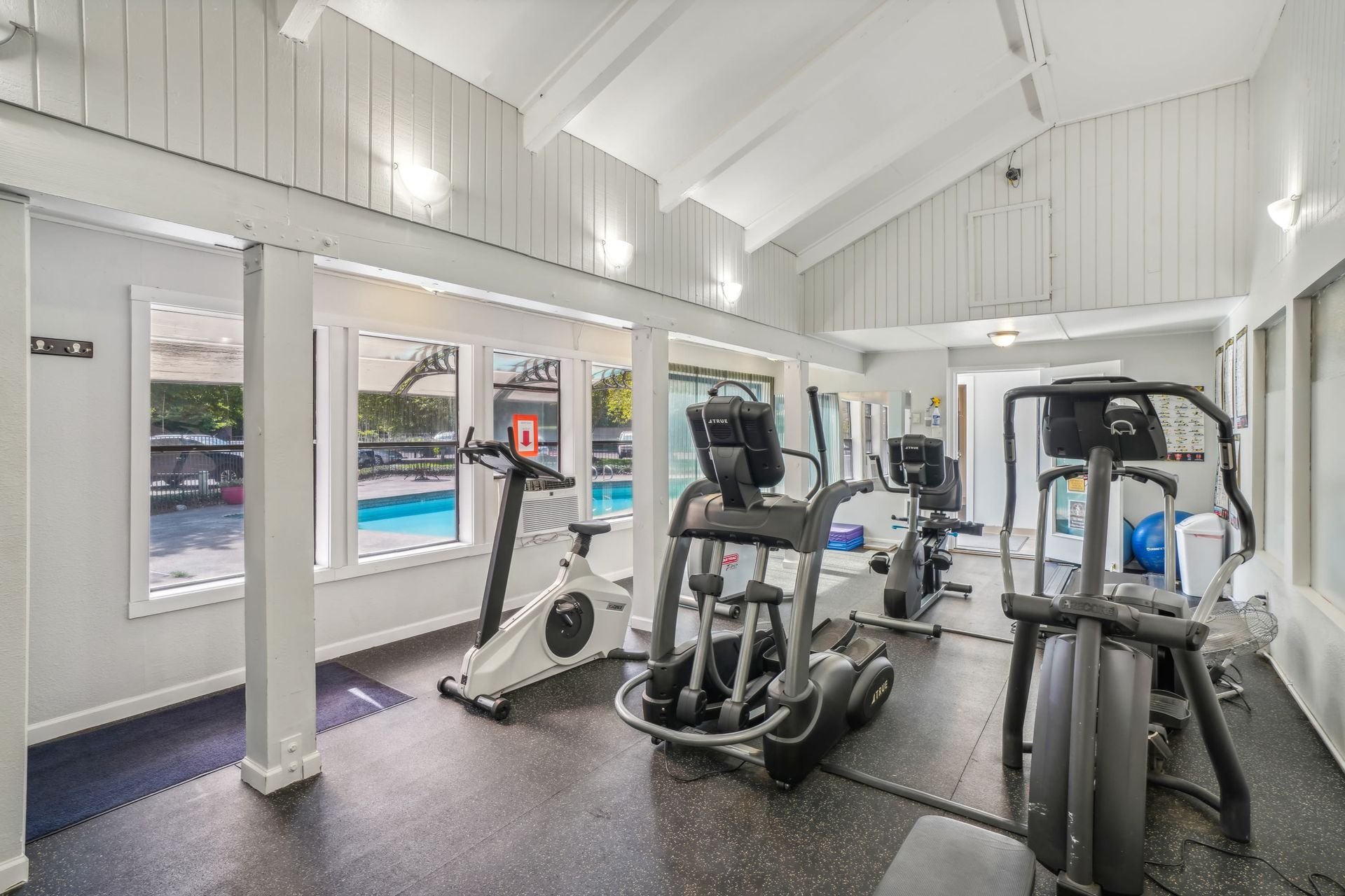 Indoor community gym with cardio machines and windows overlooking the pool.