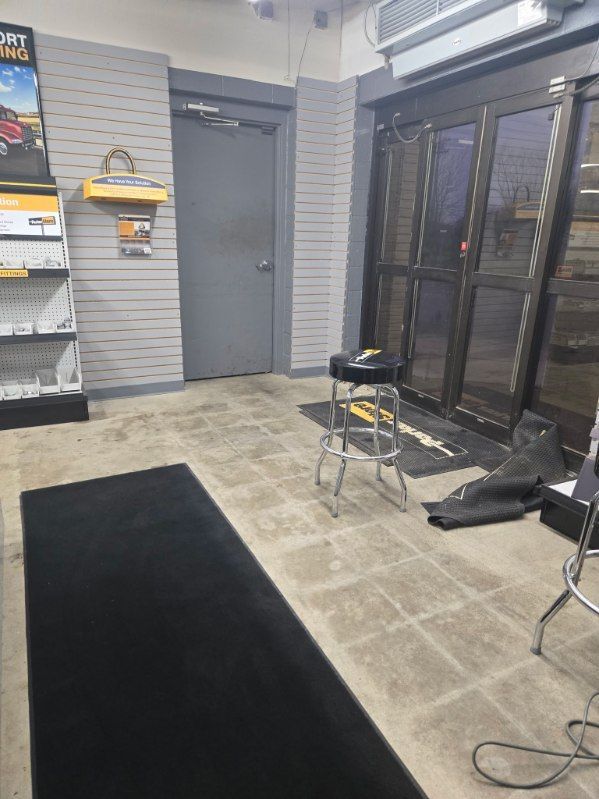 Interior of a store with a door, black mat, stool, and glass doors. Gray walls, tiled floor.