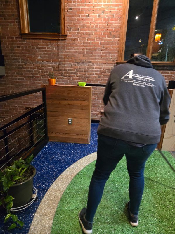 Person in gray hoodie stands on turf, looking at a wooden structure with plants and a brick wall backdrop.