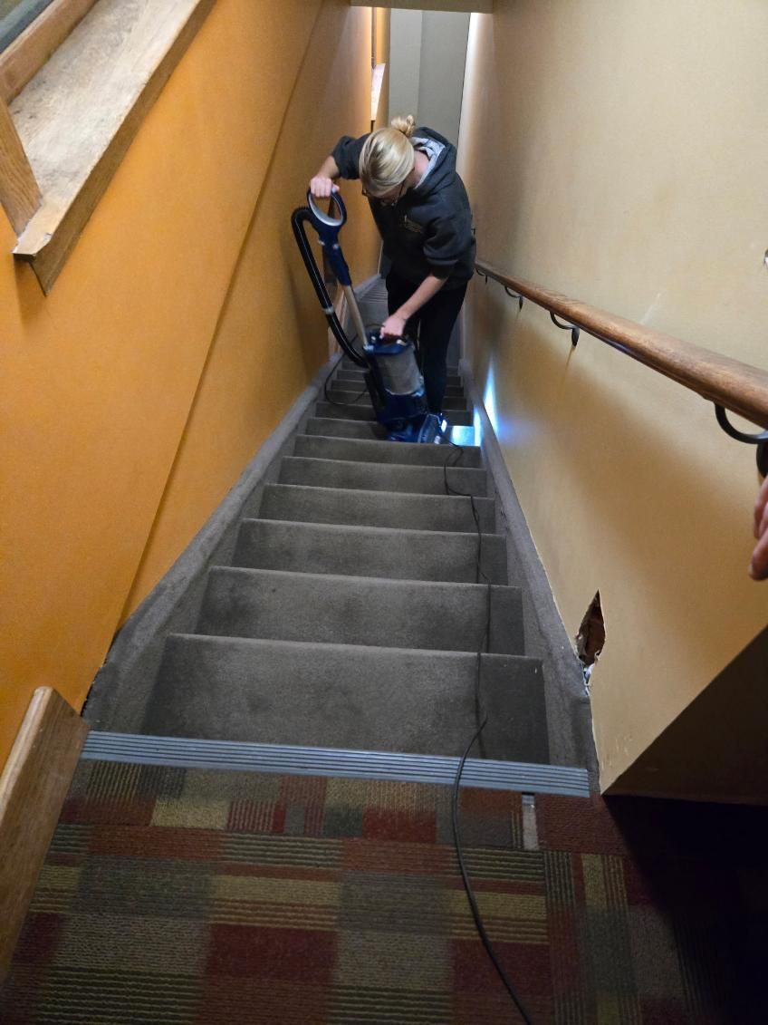 Person vacuuming a carpeted staircase with light orange walls, wooden railing, and patterned rug at the bottom.